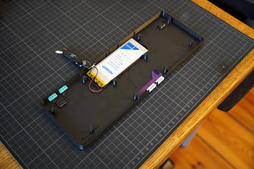 the printed base lying open on a cutting mat, with a silver LiPo battery and the purple adapter PCB mounted inside it