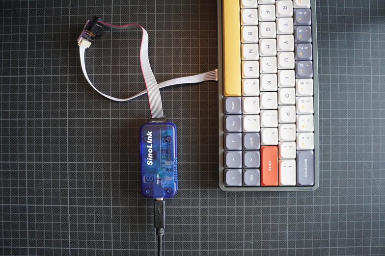 a translucent blue SinoLink programmer on a cutting mat, wired through a small adapter board and a ribbon cable into the edge of the Air60
